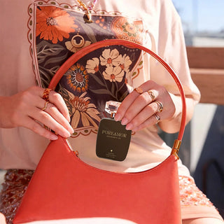 Person holding a small black bottle labeled 'PUREAMOR' with a floral-patterned handbag in the background.