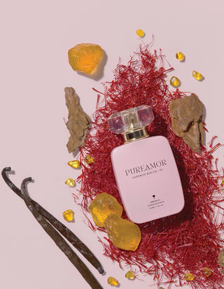 30ml Perfume bottle labeled 'PUREAMOR' surrounded by red fibers, yellow stones, and a vanilla bean on a light pink background.