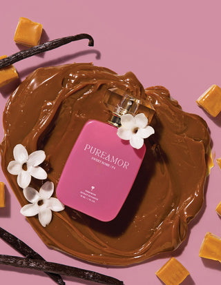 Pink 30ml perfume bottle labeled 'PUREAMOR' surrounded by chocolate, vanilla beans, and flowers on a pink background.