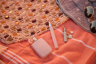 Pink perfume bottle, travel spray, and keychain on a textured orange surface with sunglasses and straw bag.