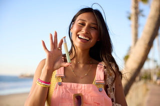 Woman on a beach holding a 12ml travel spray, wearing a pink dress and smiling.