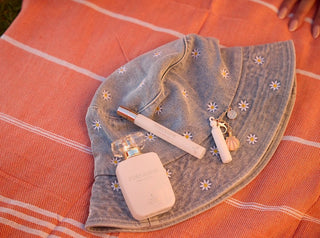 perfume bottle, travel spray, and keychain placed on a denim hat, with a relaxed summer vibe on an orange-striped fabric.