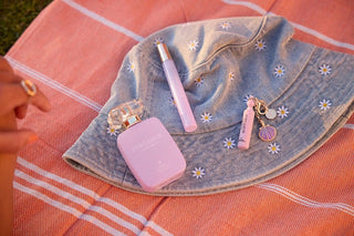 Pink perfume bottle and lip gloss on a denim bag with floral embroidery on a pink checkered tablecloth.