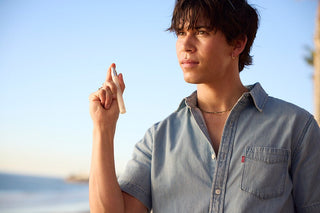 Person holding a Santal Luxe 12ml travel spray wearing a light blue denim shirt with a blurred beach background.