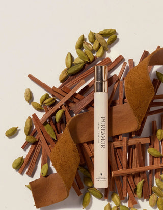PureAmor 12ml travel spray surrounded by cinnamon sticks, cardamom pods, and leaves on a beige background