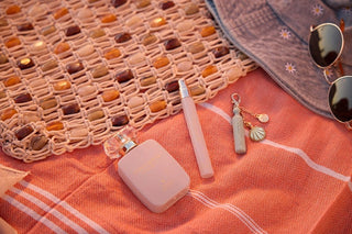 Pink perfume bottle, lip balm, and keychain on a textured orange surface with sunglasses and straw bag.